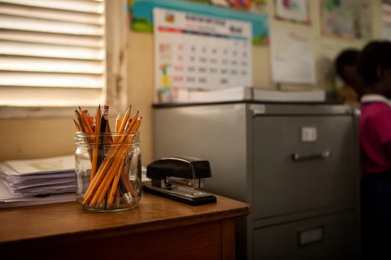 A jar of pencils and a stapler on a school office desk, a calendar blurred on the wall behind