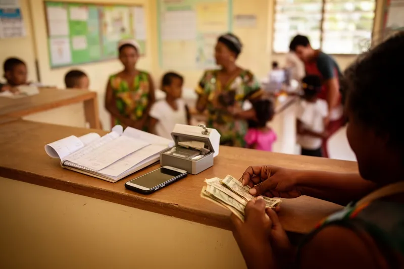 A school office counter with a receipt book, a phone and a cash box, a parent's hands at the edge of the frame