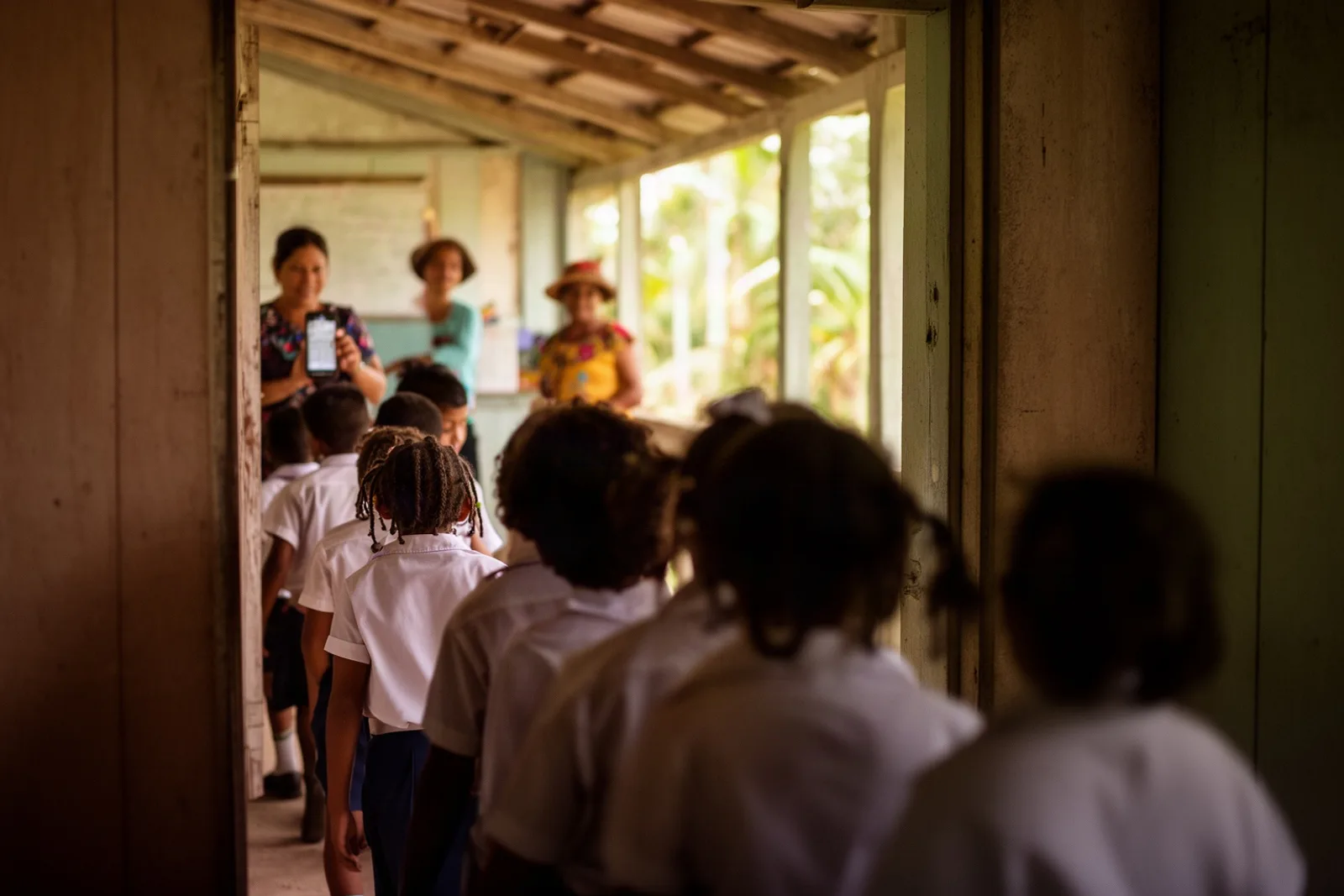 A teacher with a phone at a classroom door as children file in, seen from behind